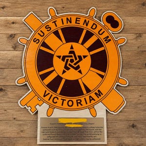 May include: Wooden plaque featuring an orange and brown emblem with the words "SUSTINENDUM VICTORIAM." The emblem includes a star, a key, and a ship's wheel design. A small plaque below contains text.