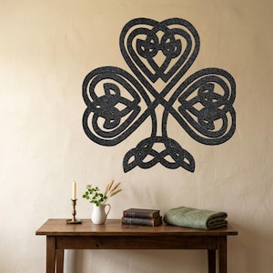 May include: A large, dark grey Celtic knot shamrock wall art. The three-leaf clover design is mounted on a light beige wall. A wooden table in the foreground holds a candle, vase, books, and a folded green blanket.