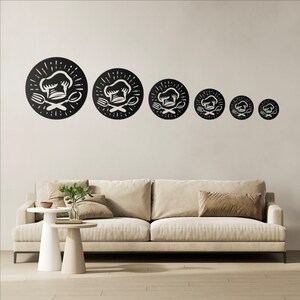 May include: Six black metal wall art pieces, each with a chef's hat, fork, and spoon design. The circular pieces are of varying sizes and are mounted on a neutral wall above a beige sofa. The design is suitable for a kitchen or dining room.