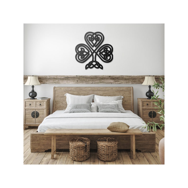 May include: A black metal shamrock wall decoration featuring a Celtic knot design. The clover-shaped artwork is mounted on a white wall above a wooden bed. The bedroom has a rustic, natural aesthetic with wooden furniture.