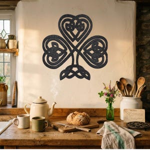May include: A dark grey Celtic knotwork shamrock wall decoration. The clover is mounted on a cream wall in a kitchen. The kitchen features a wooden countertop, a teapot, mugs, and a loaf of bread. The clover's design is highly detailed.