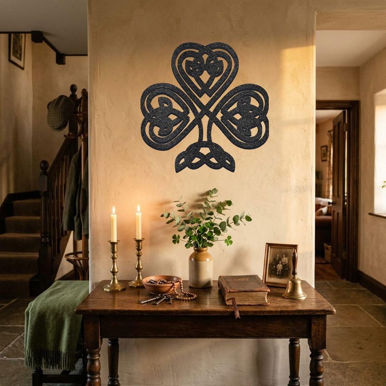 May include: A black metal shamrock wall decoration with a detailed knotwork design, mounted on a beige wall. A wooden table below holds candles, a vase of greenery, and other decorative items. The room has a rustic feel.