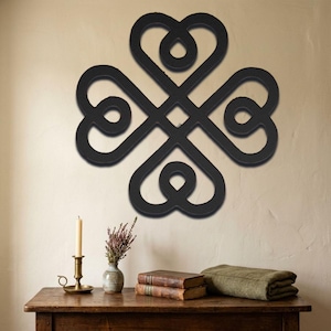 May include: Black metal wall art featuring a four-leaf clover design with heart-shaped elements. The artwork is mounted on a beige wall. A wooden table below displays a candle, books, and a vase with flowers.