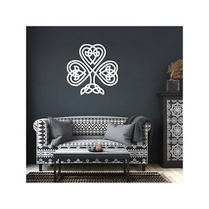 May include: A white Celtic knot shamrock wall decoration. The three-leaf clover design is mounted on a dark blue wall above a patterned sofa and a black coffee table. The wall art is a decorative element for home interiors.