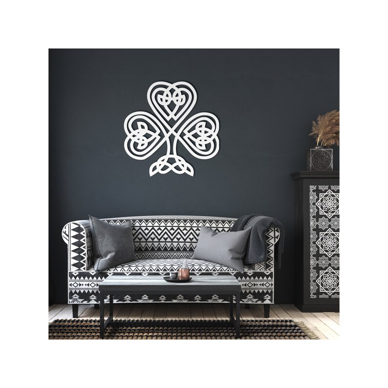 May include: A white Celtic knot shamrock wall decoration. The three-leaf clover design is mounted on a dark blue wall above a patterned sofa and a black coffee table. The wall art is a decorative element for home interiors.