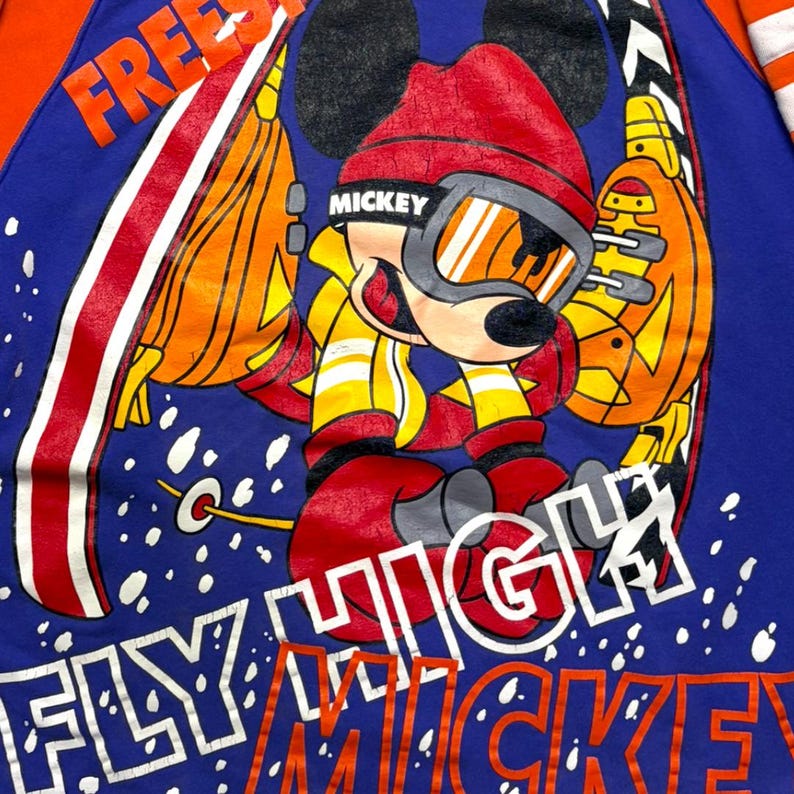 May include: A blue long-sleeve shirt featuring a graphic of Mickey Mouse wearing a red beanie and ski goggles while skiing. The design includes orange and white accents, the text "FREESTYLE" at the top, and "FLY HIGH MICKEY" at the bottom.