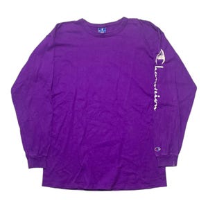 Puede incluir: Una camiseta Champion morada de manga larga. La camiseta tiene cuello redondo y el logo de Champion es visible en la manga izquierda y en el pecho. El logo de Champion también está en la etiqueta en la parte posterior del cuello.