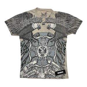 Może przedstawiać: Szary T-shirt z nadrukiem graficznym przedstawiającym dwa orły trzymające tarczę z krzyżem i tekstem "Liberias Justitia" w stylu gotyckim. Teksty "Mafia" i "Lugermuns" znajdują się na banerach pod tarczą. Tekst "Seventeen" znajduje się na banerze pod krzyżem. Tekst "Weirdo" znajduje się na białej naklejce w prawym dolnym rogu koszulki.