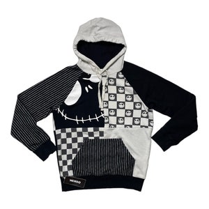 May include: Black and white patchwork hoodie with a Jack Skellington face design. The hoodie has a checkered pattern, stripes, and a black and white smiley face pattern. The hoodie has a large pocket on the front and the word "WEIRDO" is printed on a tag.