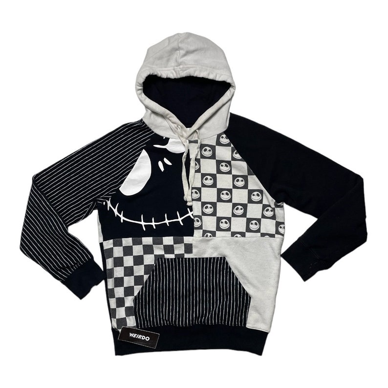 May include: Black and white patchwork hoodie with a Jack Skellington face design. The hoodie has a checkered pattern, stripes, and a black and white smiley face pattern. The hoodie has a large pocket on the front and the word "WEIRDO" is printed on a tag.