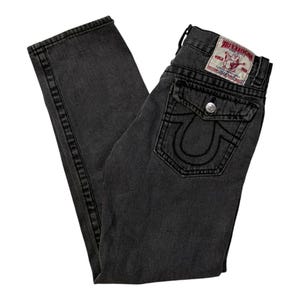 May include: Charcoal grey denim jeans featuring a flap pocket on the back with a silver button and a signature horseshoe stitch design. A white True Religion brand patch with red text and a Buddha logo is stitched onto the waistband above the pocket.