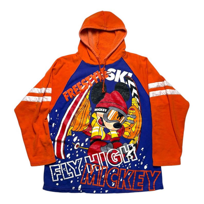 May include: An orange and blue hooded sweatshirt featuring a graphic of Mickey Mouse wearing ski gear. The text "FREESTYLE SKI" and "FLY HIGH MICKEY" is printed on the front. The sleeves have two white horizontal stripes, and the hood has drawstrings.