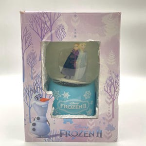 Puede incluir: Una bola de nieve de Frozen II en caja con Elsa y Anna en el interior. La base es azul claro con el logotipo de Frozen II. La caja es lavanda con diseños de árboles y hojas, y una imagen de Olaf el muñeco de nieve.