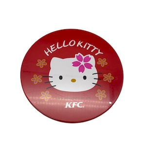 Puede incluir: Una lata metálica circular roja que presenta la cara del personaje Hello Kitty con una flor rosa en la cabeza. El texto HELLO KITTY está impreso en blanco sobre la cara y el logotipo de KFC está impreso en blanco en la parte inferior.