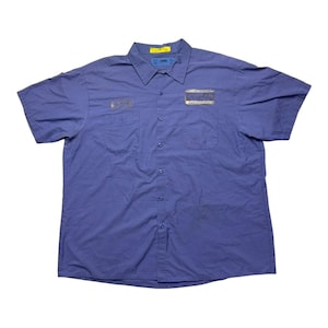 May include: A short-sleeved button-down shirt in a solid periwinkle blue color. The shirt features a classic collar, a front button placket, and two chest pockets. A rectangular patch is sewn above the left chest pocket, and a yellow tag is visible.
