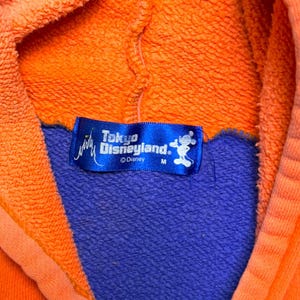 May include: A bright orange hooded sweatshirt featuring a blue interior lining and a blue rectangular tag at the neckline. The tag displays the white text Tokyo Disneyland, a small castle icon, a Mickey Mouse silhouette, and the size letter M.