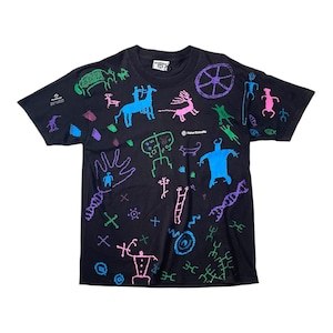 Op de afbeelding: Zwart t-shirt met een kleurrijke all-over print van prehistorische figuren en symbolen. Het ontwerp bevat dieren, mensachtige figuren en geometrische vormen in blauw, groen, roze en paars. Het shirt heeft een ronde hals en korte mouwen.