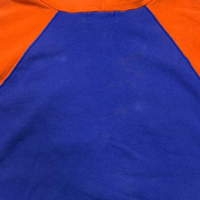 May include: A long-sleeve shirt featuring a vibrant blue central panel and bright orange raglan sleeves. The garment has a round neckline and visible stitching along the diagonal seams where the orange sleeves meet the blue body of the shirt.