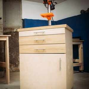 Puede incluir: Un banco de trabajo de madera con tres cajones y una puerta de armario. El banco de trabajo tiene ruedas negras y una taladradora de columna montada en la parte superior. La taladradora de columna es naranja y negra.