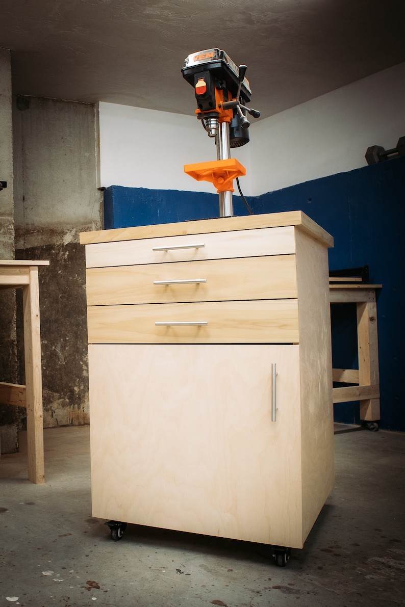 Puede incluir: Un banco de trabajo de madera con tres cajones y una puerta de armario. El banco de trabajo tiene ruedas negras y una taladradora de columna montada en la parte superior. La taladradora de columna es naranja y negra.
