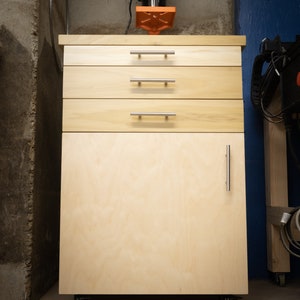 Puede incluir: Un armario de madera con tres cajones y una puerta sobre ruedas. El armario est&aacute; hecho de madera clara y tiene tiradores plateados. Una taladradora de columna negra est&aacute; colocada encima del armario.