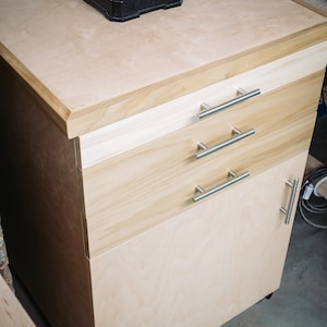 Puede incluir: Un armario de madera clara con tres cajones y una puerta. El armario tiene tiradores de metal plateado y est&aacute; sobre ruedas. La parte superior del armario est&aacute; hecha de madera contrachapada y tiene un objeto de metal negro en la parte superior.