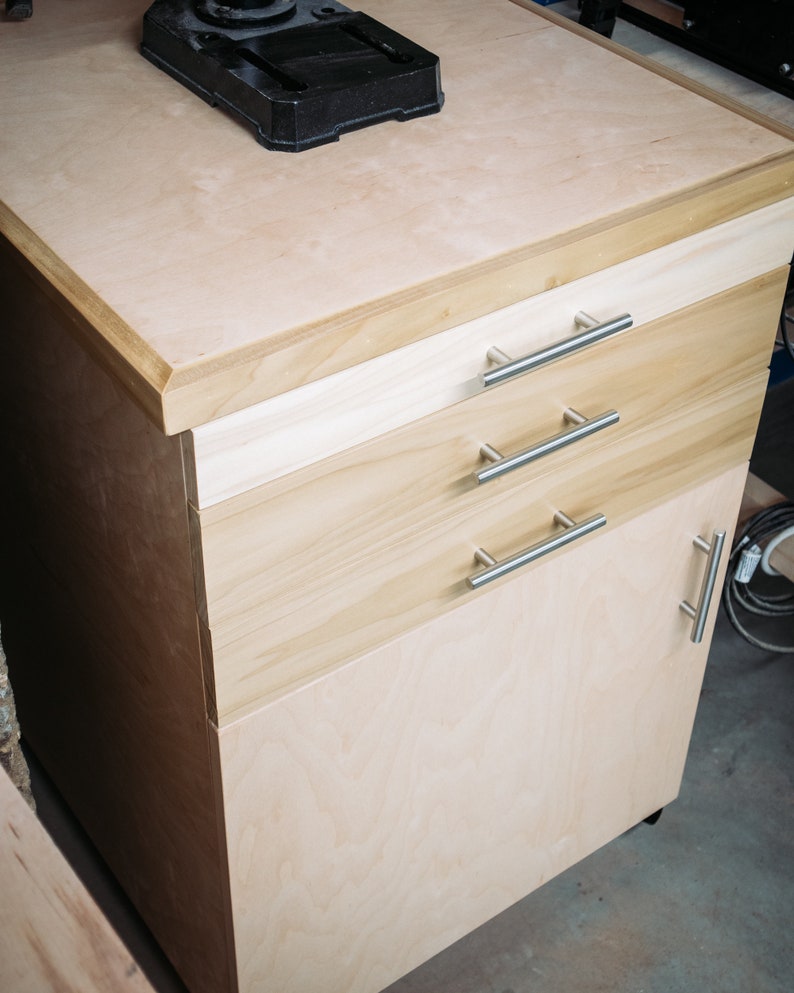 Puede incluir: Un armario de madera clara con tres cajones y una puerta. El armario tiene tiradores de metal plateado y est&aacute; sobre ruedas. La parte superior del armario est&aacute; hecha de madera contrachapada y tiene un objeto de metal negro en la parte superior.
