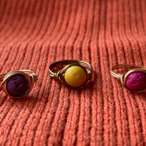 Puede incluir: Tres anillos de alambre dorado con cuentas de roca volcánica de colores. Los anillos están sobre una superficie naranja texturizada. Las cuentas son moradas, amarillas y rosadas.