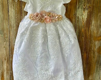 May include: White lace dress with a peach and ivory flower sash. The dress has short sleeves and a scalloped hem.
