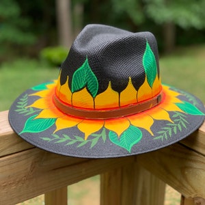 May include: A black straw hat with a painted sunflower design. The sunflower has yellow petals, orange center, and green leaves. The hat has a brown leather band.