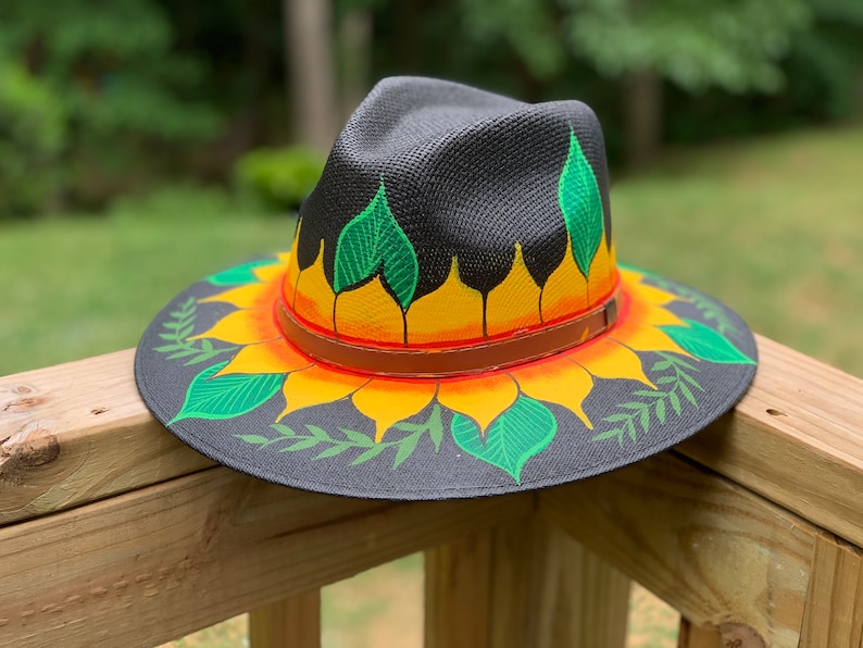 May include: A black straw hat with a painted sunflower design. The sunflower has yellow petals, orange center, and green leaves. The hat has a brown leather band.