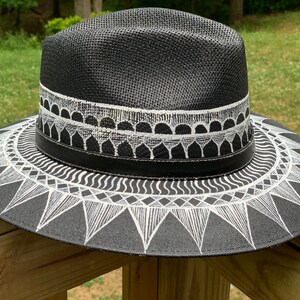 May include: A black straw hat with a white geometric design painted on the brim. The design features a series of triangles, waves, and circles.