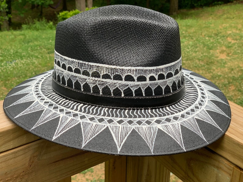 May include: A black straw hat with a white geometric design painted on the brim. The design features a series of triangles, waves, and circles.