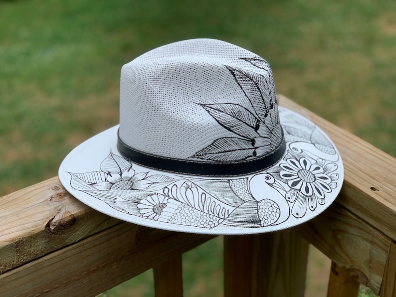May include: A white straw hat with a black band and black ink drawings of birds, flowers, and leaves.