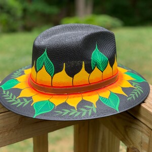 May include: A black straw hat with a painted sunflower design. The sunflower has yellow petals, orange centers, and green leaves. The hat has a brown leather band.