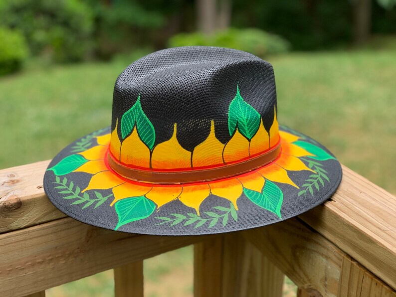 May include: A black straw hat with a painted sunflower design. The sunflower has yellow petals, orange centers, and green leaves. The hat has a brown leather band.