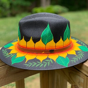 May include: A black straw hat with a painted sunflower design. The sunflower has yellow petals, orange center, and green leaves. The hat has a brown leather band.