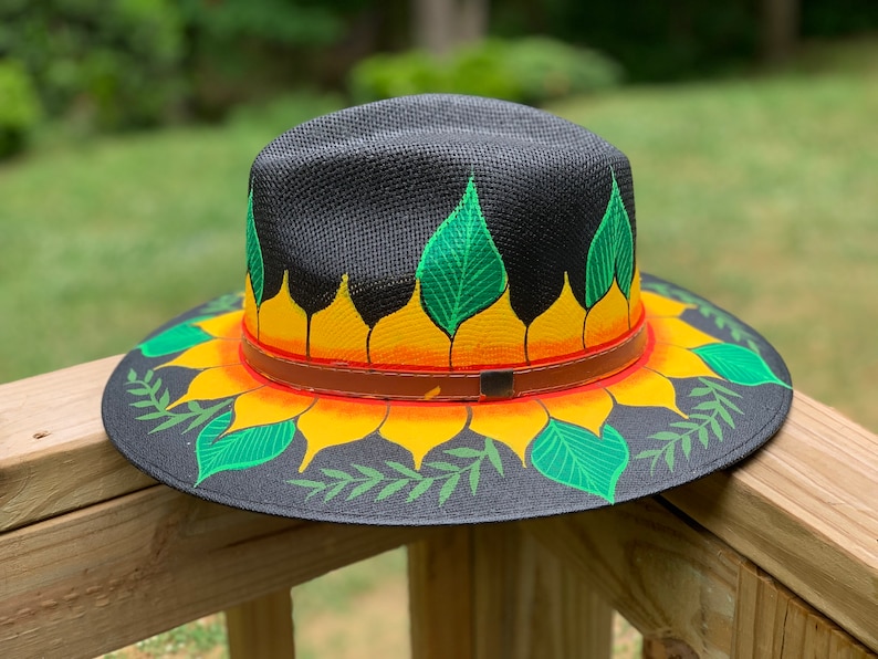 May include: A black straw hat with a painted sunflower design. The sunflower has yellow petals, orange center, and green leaves. The hat has a brown leather band.