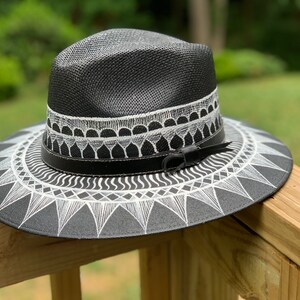 May include: A black straw hat with a white geometric design painted on the brim. The hat has a black leather band with a buckle.