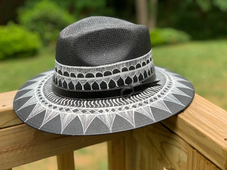 May include: A black straw hat with a white geometric design painted on the brim. The hat has a black leather band with a buckle.