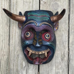 Pode incluir: Uma máscara de madeira preta com chifres, pintada de vermelho, verde, dourado e azul. A máscara tem uma boca larga com dentes afiados e uma língua vermelha que se projeta.