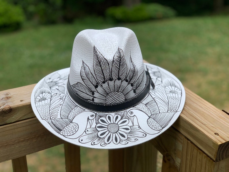 May include: A white straw hat with a black leather band and black ink drawings of flowers, birds, and a sun on the brim.