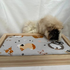 May include: A wooden bed for small pets, with a gray and white patterned fabric liner. A long-haired guinea pig is lying on the bed.