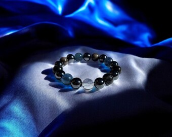 May include: A beaded bracelet with black and blue stones and a clear crystal charm. The bracelet is lying on a blue satin fabric.
