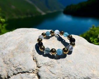May include: A bracelet made of black, gold, clear, and blue beads. The bracelet is on a gray rock with a blue lake and green mountains in the background.