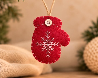 May include: A red felt mitten ornament featuring white embroidered snowflake detailing and a small light-colored wooden button at the top. The mitten is suspended by a thin, natural-colored jute twine loop for hanging.