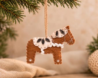 May include: A brown felt horse ornament featuring a cream-colored saddle and mane with dark gray leaf-shaped accents. The horse is decorated with white stitched details and small star patterns on its legs, suspended by a thin jute twine loop.