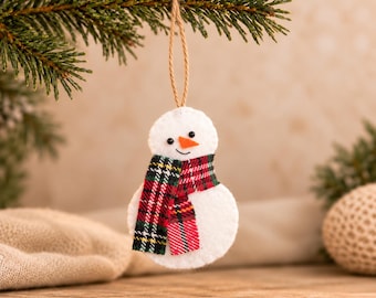May include: A white felt snowman ornament featuring a small orange felt nose, two black bead eyes, and a stitched smile. The snowman wears a red, green, and black plaid fabric scarf and hangs from a thin, light brown jute twine loop.