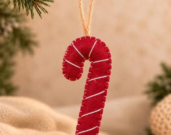 May include: A red felt candy cane ornament featuring white stitched diagonal stripes and a blanket-stitched edge. The ornament hangs from a natural jute twine loop, showcasing a soft, handcrafted texture with visible thread details.