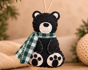 May include: A black felt bear ornament with a cream-colored muzzle and paw pads. The bear features white whip-stitching along the edges and wears a green and white gingham scarf. A tan twine loop is attached to the top for hanging purposes.